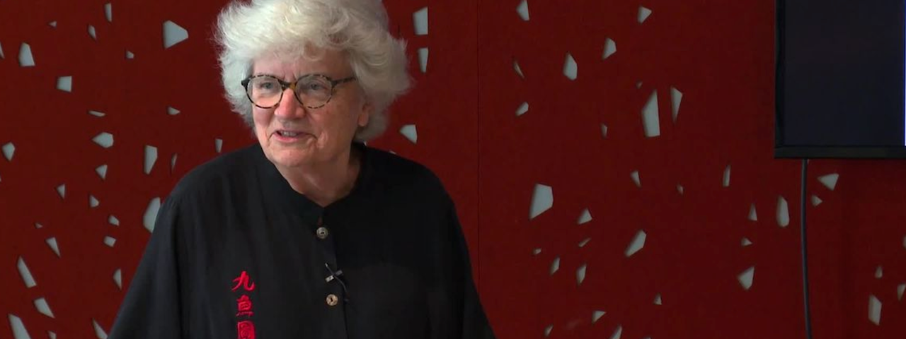 Photo of an older woman wearing glasses and a clip-on microphone, speaking behind a podium.