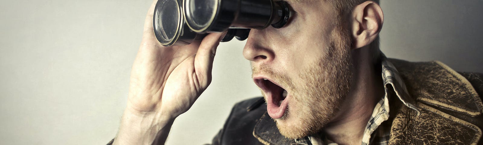 A blonde guy in his 40s wearing a leather jacket looks through binoculars and gasps