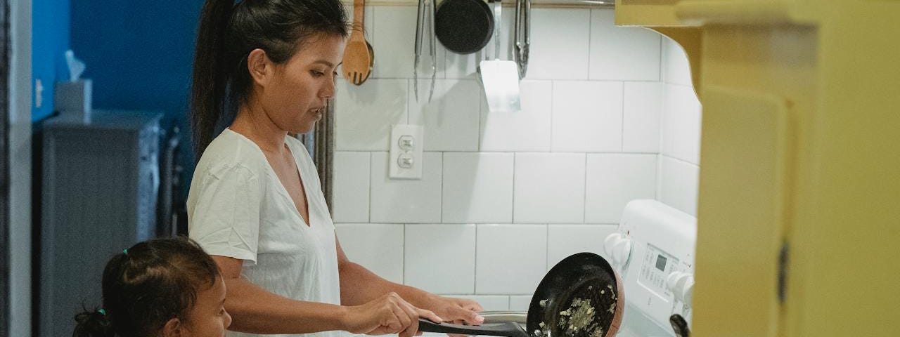 Seorang perempuan Asia yang memasak sambil menjaga anak kecil. An asian female cooking while taking care of the child in a kitchen.