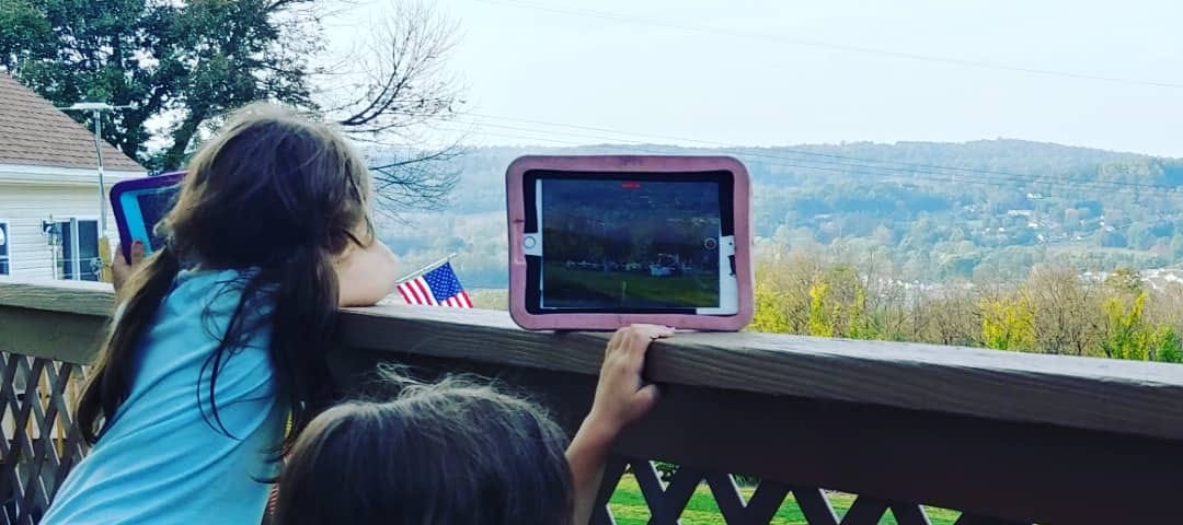 Two young girls sitting on a brown deck looking over the railing and holding their iPads on the railing