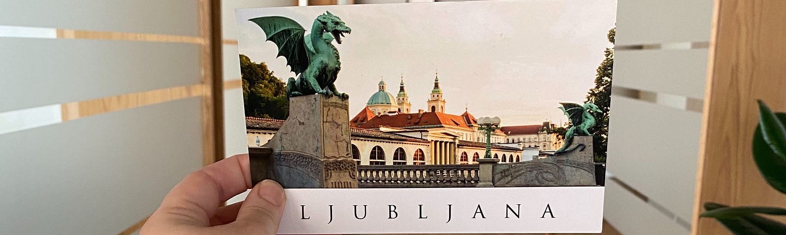 An arm holding a postcard of a dragon statue and building, the postcard reads, “Ljubljana.”
