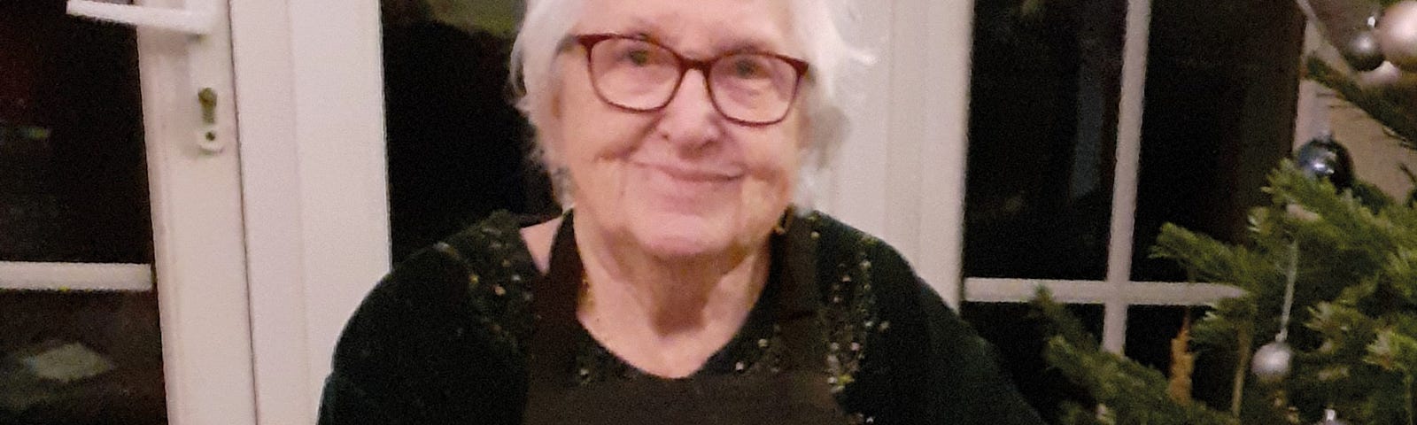 An older woman, Jackie, smiles at the camera while holding a gift sack. Next to her is a Christmas tree.