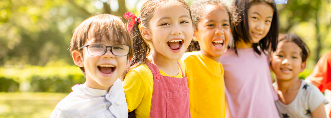 Five elementary school-aged children stand outside, smiling with their arms around each other.