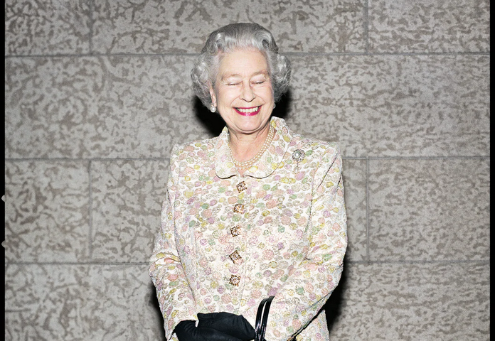 The late Queen Elizabeth the second standing in the center of the frame. She’s wearing a yellow floral print jacket and skirt. Wearing black gloves with a black purse hanging from her left wrist. Her eyes are closed and she’s smiling with lips painted red. It’s not a regular boring image of her because of the look on her face. Photograph by Christopher Wahl.