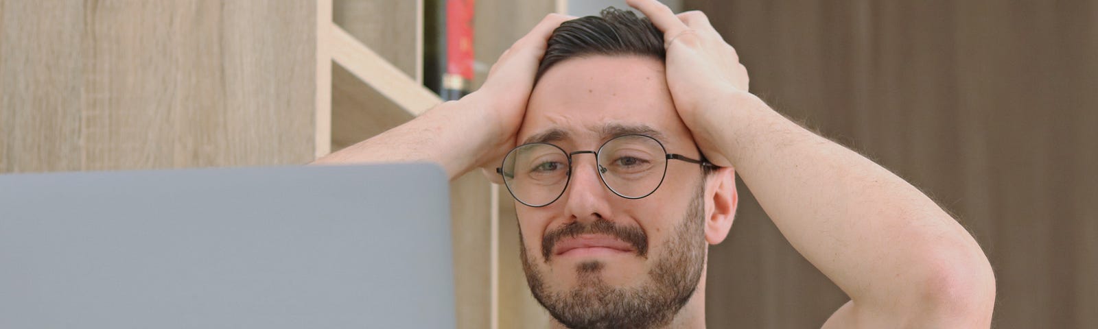 A man in front of his laptop, worried, holding his head with both hands.