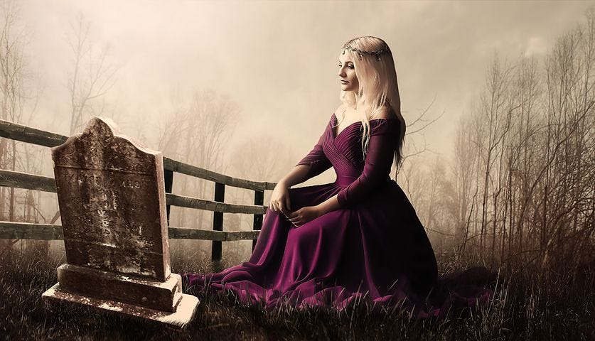 A beautiful girl, wearing a gorgeous maroon dress, sitting in front of an old grave’s  headstone.