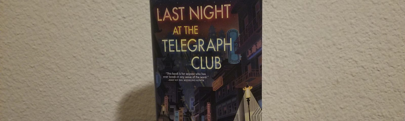 The book stands vertically atop a dark brown wooden tabletop with a white wall behind it. The title is displayed at the top in large letters. Below it is a cityscape that looks like Chinatown. Underneath a street lamp on the right side is the main character, Lily Hu, and the love interest standing in front of her within the shadows of the alley. The author’s name is in the bottom left corner.