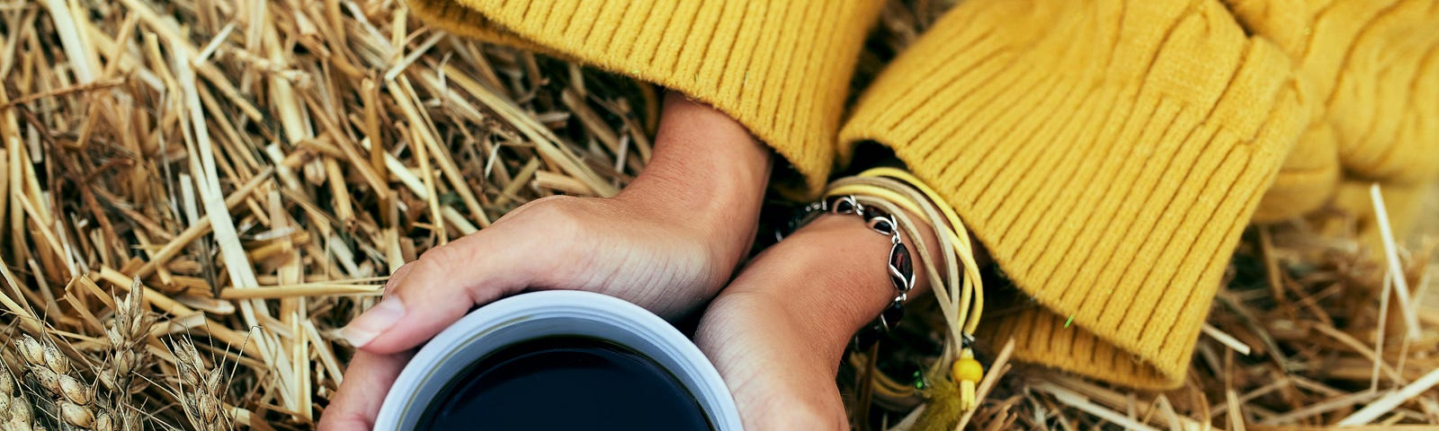 A woman in a yellow sweater holds a cup of coffee in both hands, with stalks of grain beneath it.
