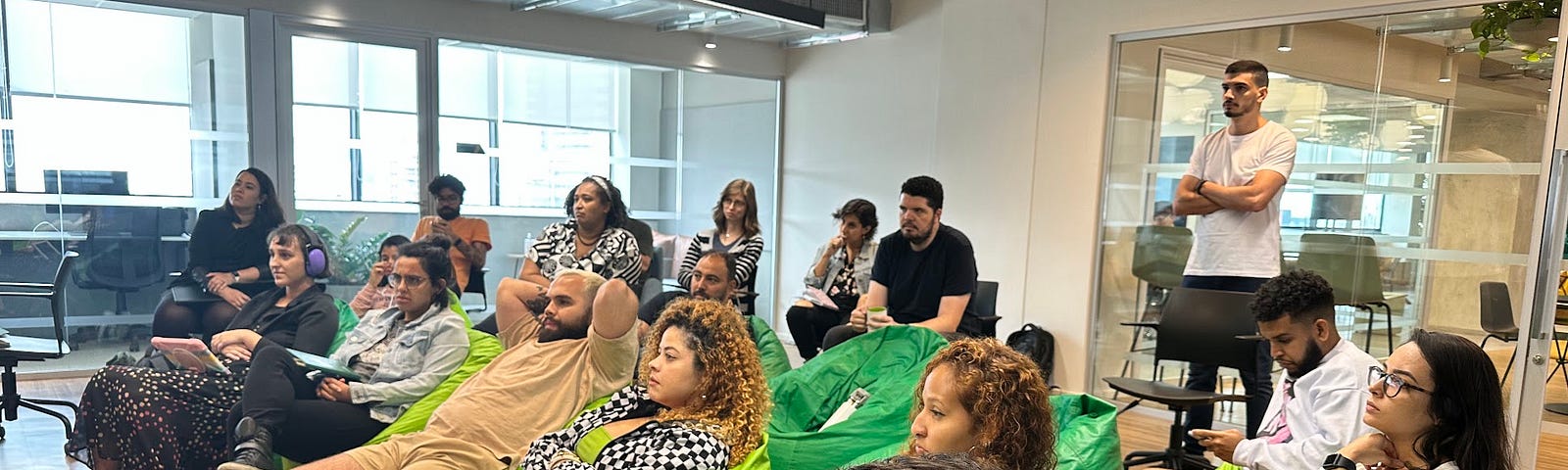 Foto que mostra várias pessoas no ambiente interno do Hub do Jusbrasil em São Paulo, sentadas em puffs verdes. Eu apareço à esquerda, usando abafadores de ruído roxos e com um tablet no colo.