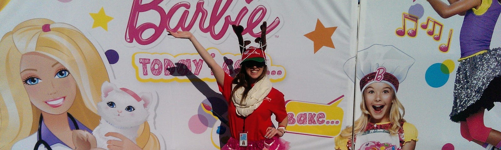 A girl in a red shirt with a reindeer hat, scarf, and tutu stands in front of a Barbie poster.