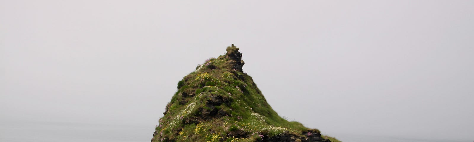 um pico de montanha de pedras, verde por cima. observa-se o mar ao fundo