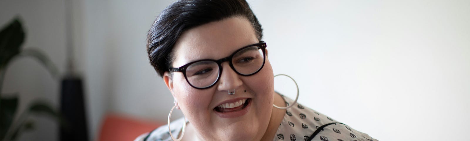 A smiling fat woman with ear and nose piercings wearing glasses.
