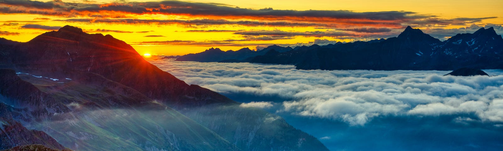 Brightly colored picture of the sun rising above mountain peaks and clouds. The sky is golden and there is a green field on top of the mountain in the foreground.