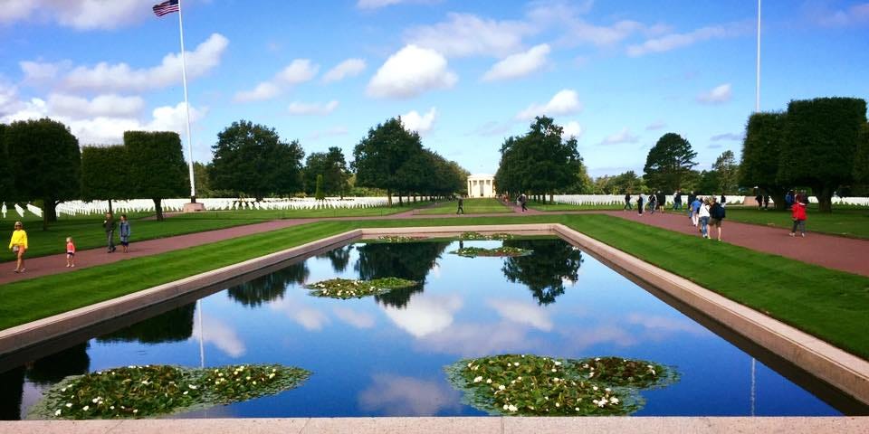 Color photo of the American Cemetery in Normandy, showing a pool reflecting the brilliant blue sky, with deep green grass surrounding the pool and white tombstones in the distance.