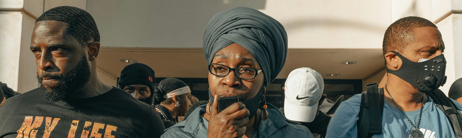 A Black woman speaks at a rally with two Black men on either side of her. Several other Black people stand behind the trio.