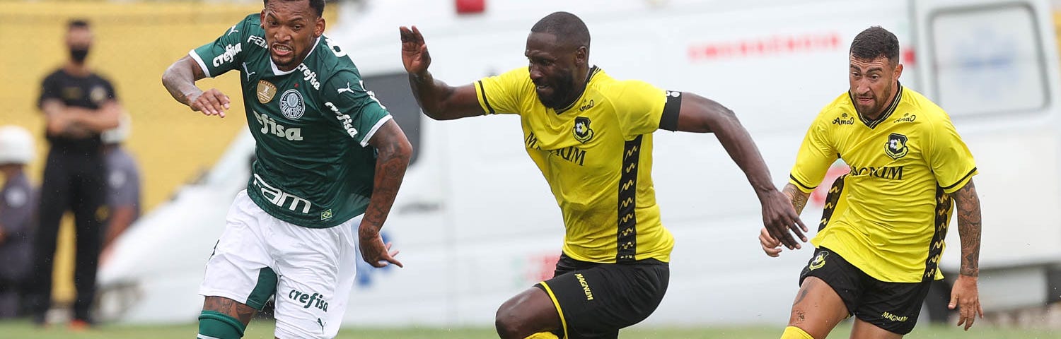 Jailson, do Palmeiras, disputa bola com o jogador Rodrigo Souza, do São Bernardo, durante partida válida pela segunda rodada do Campeonato Paulista. Os dois são negros. Jaíson veste camisa e meias verdes e calção branco. Rodrigo usa camisa e meias amarelas e calção preto.