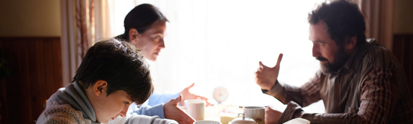 A mother and father argue in the background while their son looks down in fear at his breakfast.