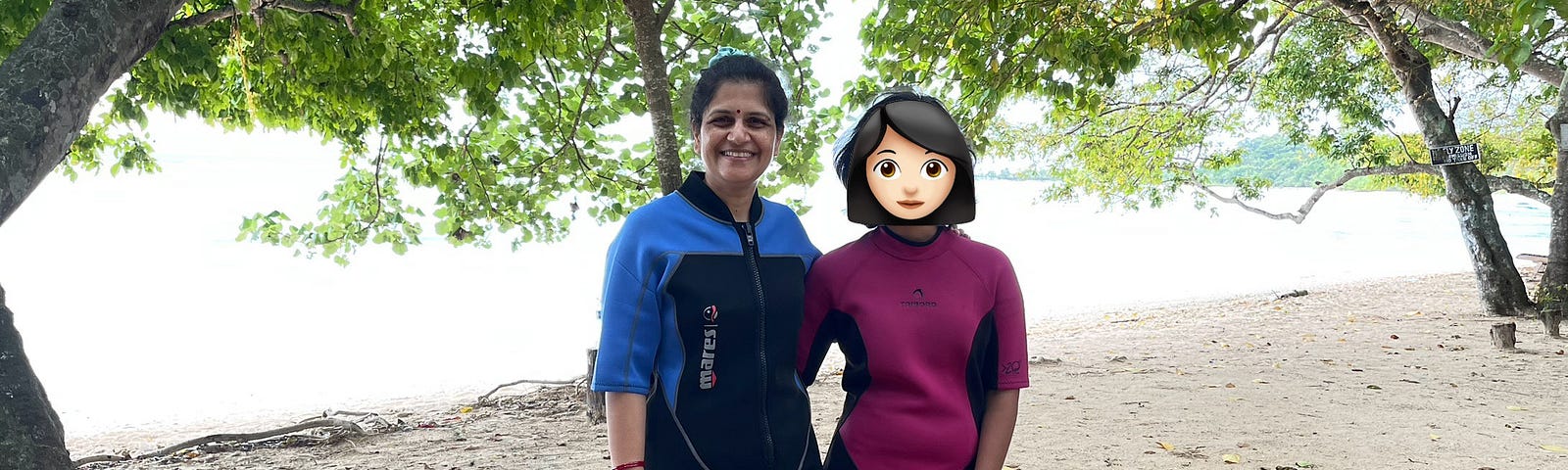 A woman and her daughter in scuba wear stand on the beach. There are green leaves on the trees on either side of them. This is a beach in a fertile place. The daughter is as tall as the mother.