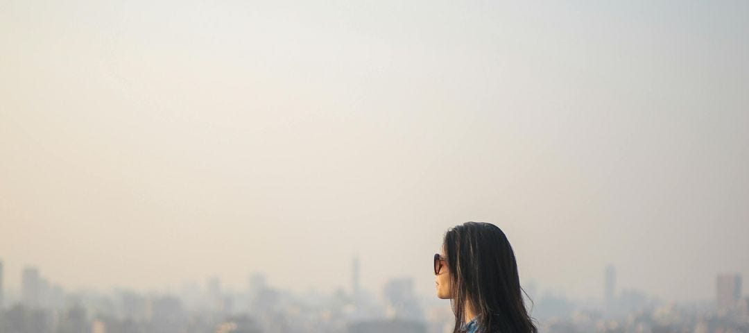 A dark-haired woman in sunglasses, a jean jacket, and a black-and-white striped skirt sits on the edge of a tall building and looks out at a hazy cityscape.