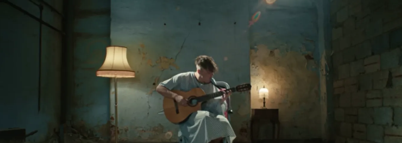 A young man in a hospital gown sits in a wheelchair in a emptied out wreck of a room, playing a guitar.