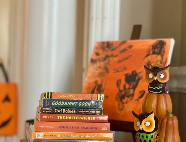 Stack of children’s Halloween books with owls figurine to the right and an orange canvas with brown handprints in the background.