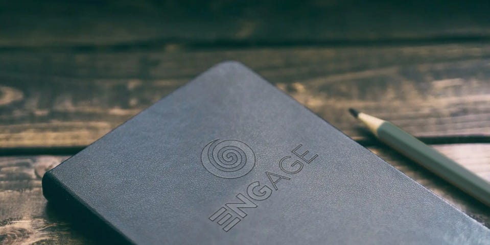 A black notebook with the Engage logo on the cover next to a pencil on a wooden table.