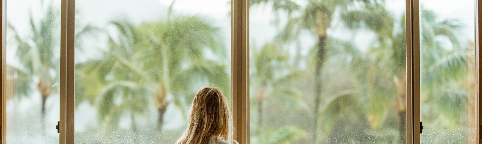 Photo of thin, blonde woman, long hair, perched at window pantless, staring at a rainy landscape of palms in fancy suite.