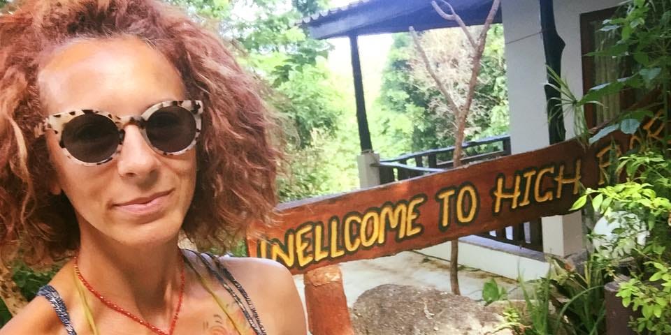 Author with big red hair, wearing shades and a spagetti-strapped dress in front of a sign that reads, “Welcome to High Bar.”