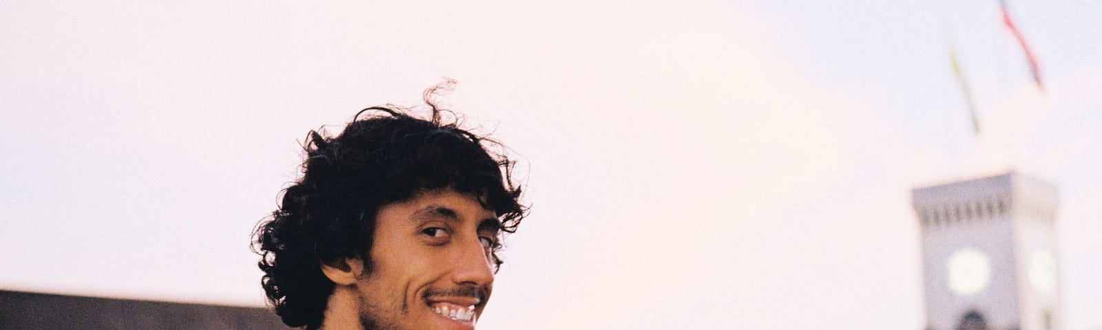 Bernardo Picão smiles at the camera looking over his shoulder. An out-of-focus clock tower is seen the background.