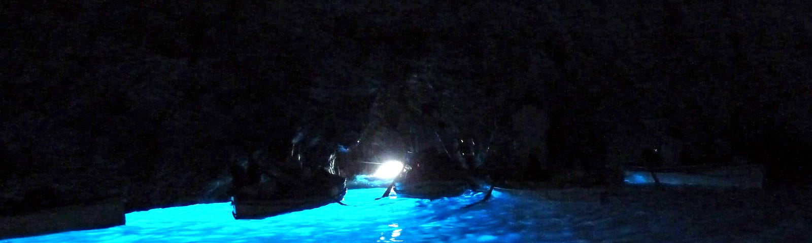 A dark cave with a small point of light in the center behind two rowboats. The water is glowing cerulean blue