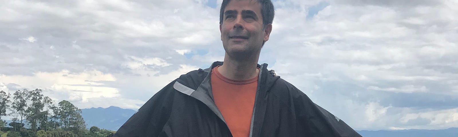 Author looking up, to the right and the distance on a pleasant, slightly cloudy day. His hands are on wooden railings behind him.