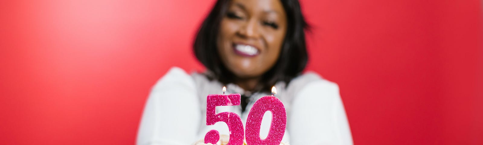A Black woman with shoulder length straight hair and a party hat wears a white jumper with a pink number 50 on the front. She is smiling and holding a birthday cake, against a red backdrop