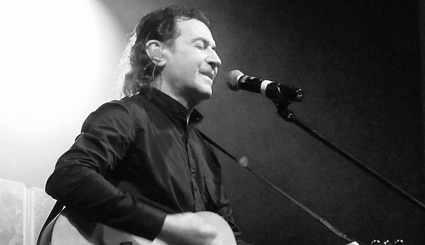 Black and white photo of Albert Hammond singing and playing an acoustic guitar.