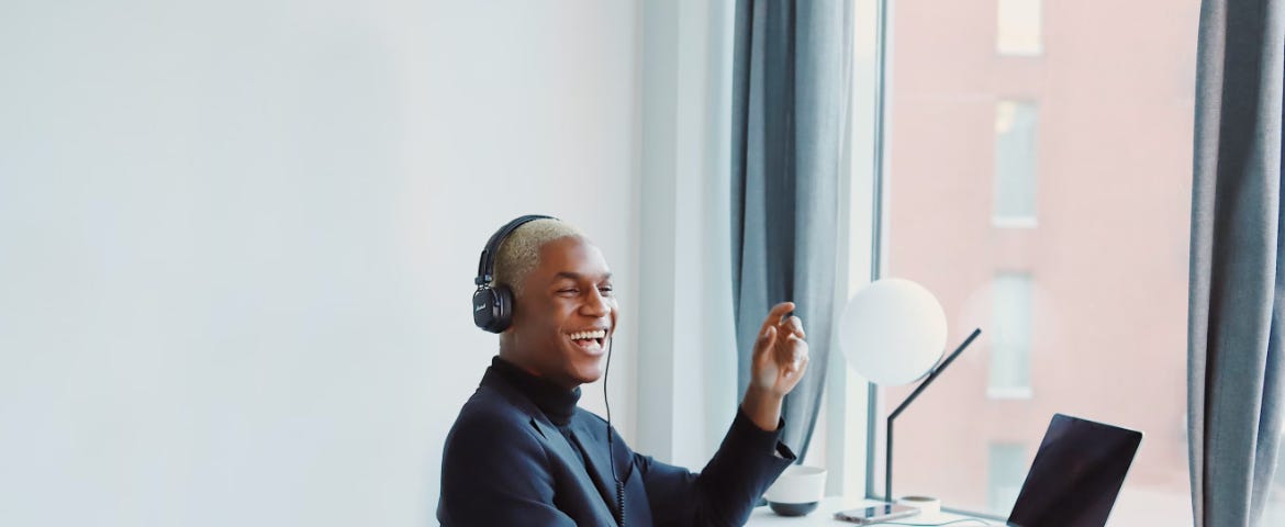 Photo of a laughing adult wearing casual clothes and headphone, seated at a desk with an open laptop.