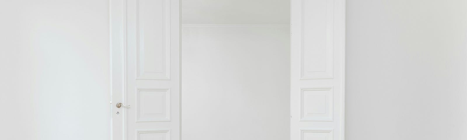 White walls in a starkly white room, with the door open looking into another white room. Clean-looking tan floorboards are the only color.