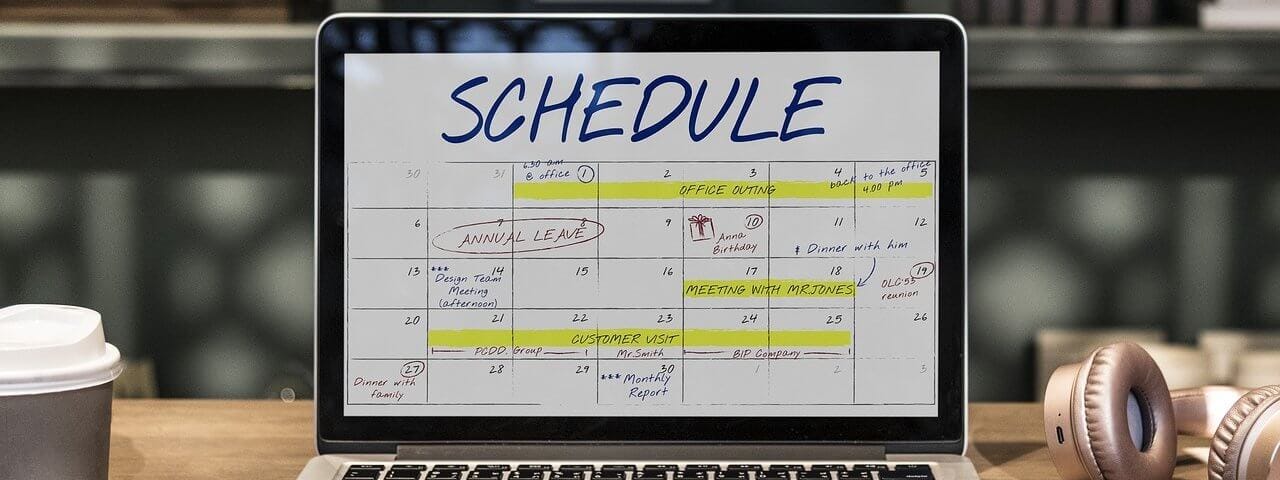 Macbook pro aberto com SCHEDULE (cronograma) escrito grande na tela em azul. Uma agenda em formato de calendário mensal abaixo com alguns dias grifados com marca texto amarelo, outros dias com círculos vermelhos marcados e outros dias com algo escrito bem pequeno em preto. O computador está sobre uma mesa de madeira com uma parte de um fone de ouvido rose gold do lado direito, assim como, um pedacinho de um celular também do lado direito.