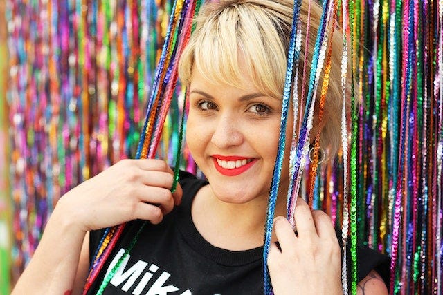 A blonde woman leaning against a wall of sequin ropes.
