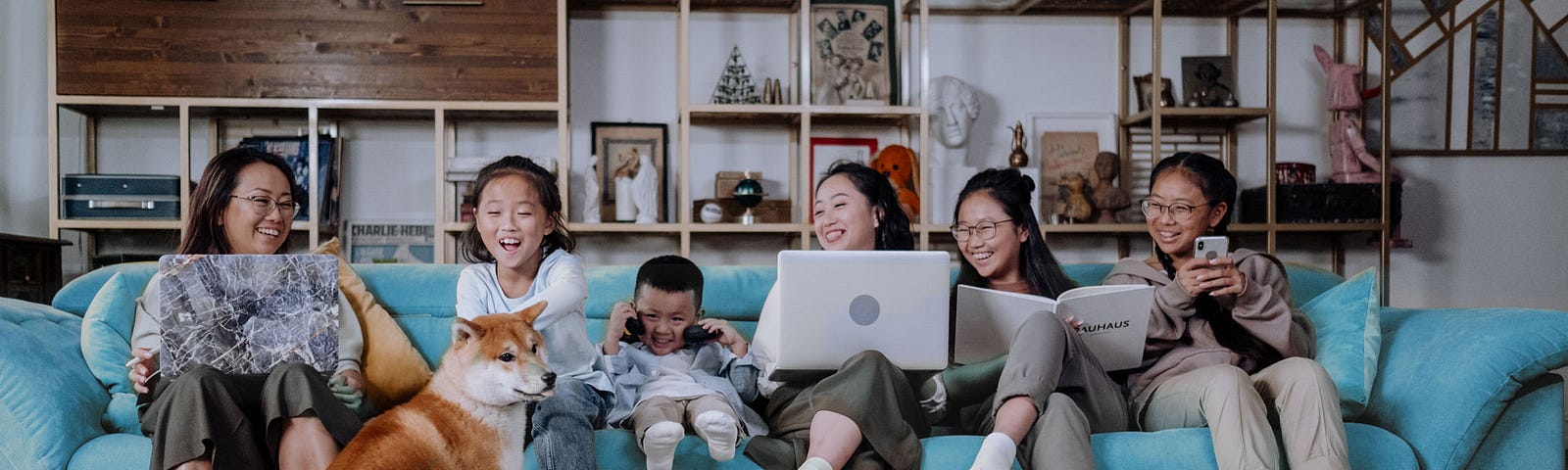 A family of six sit on a long couch on various internet enabled devices. They laugh at a dog on the floor.