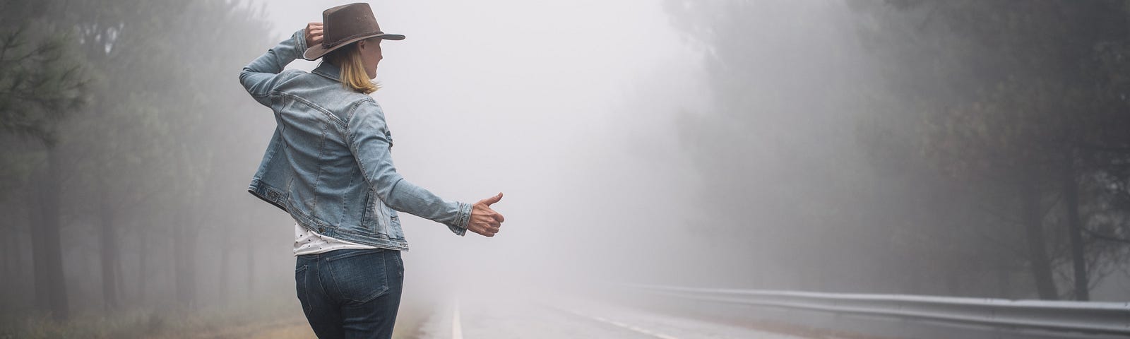 A female hitchhiker with a cowboy hat stands beside the road, her right arm extended with her thumb up.