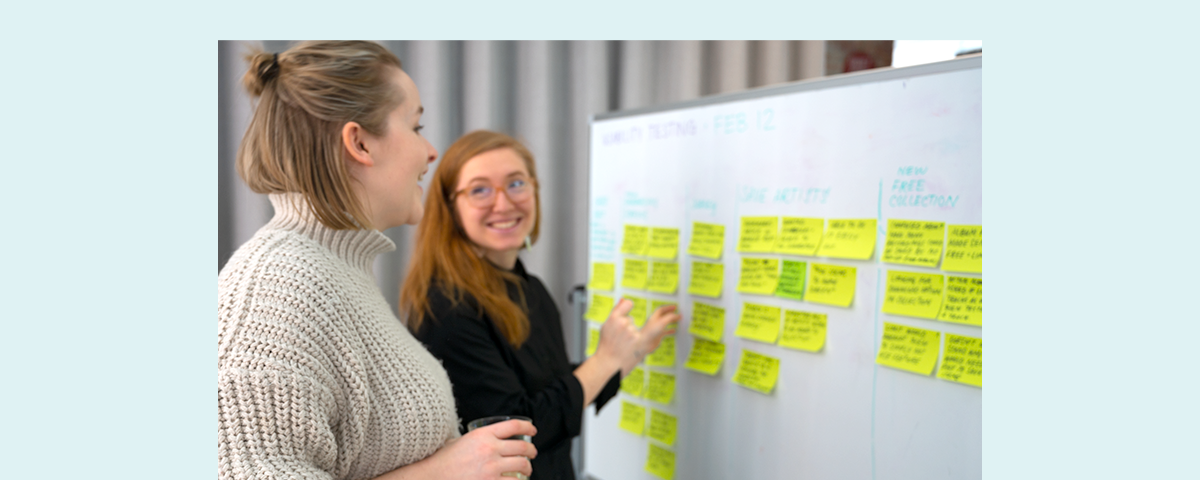 Two happy women discussing a whiteboard that is covered in sticky notes.