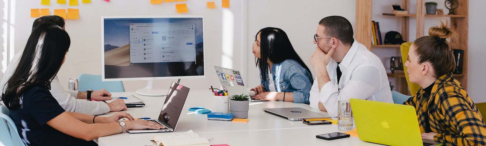 Photo of five employees sitting around a table with their laptops, looking at a computer monitor in the center. Post-it notes are on the wall behind them.