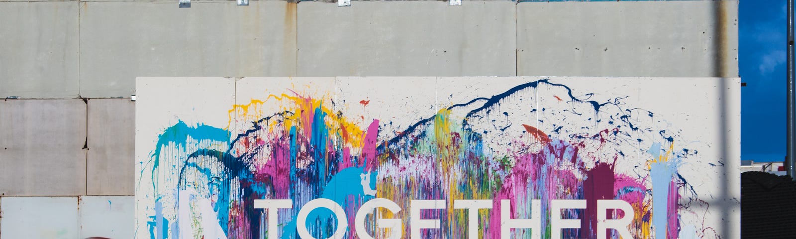 a concrete wall painted white on the lower side , splashed with colours like pink blue purple yellow and green. Written in block letters in white colour “together” on it.