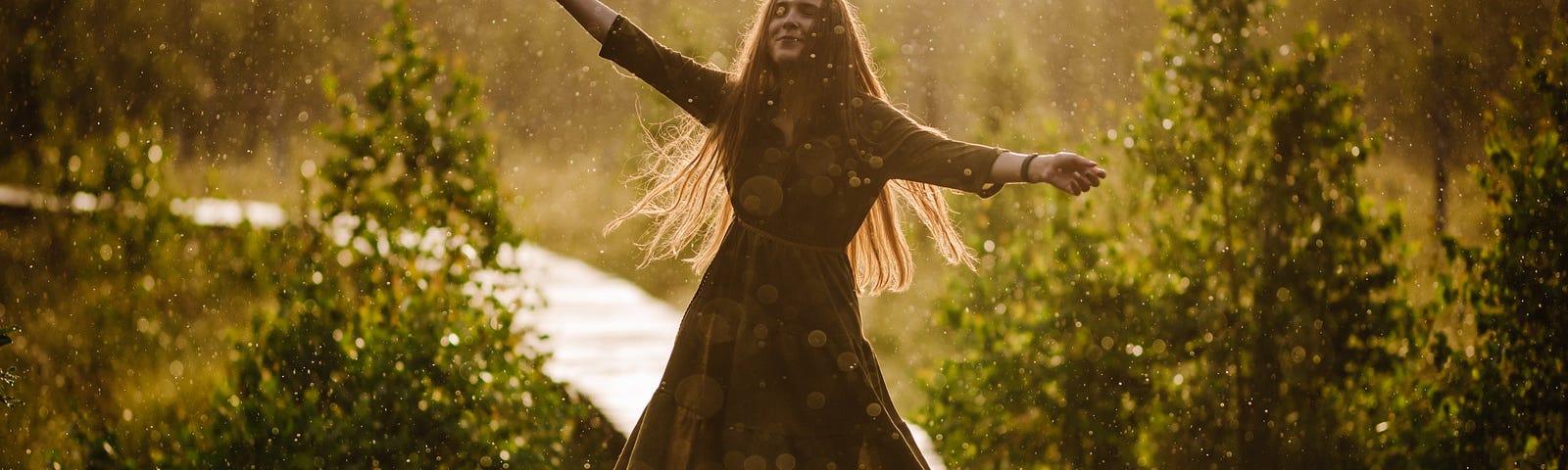 A woman dances in a spring rain-shower with a smile on her face and her arms spread wide. The sun shines through the rain.