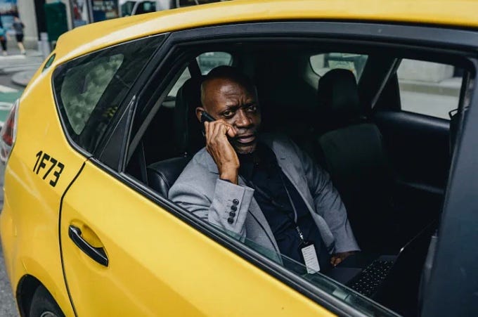 Black man wearing a suit talking on his cell phone while riding in the back seat of a yellow taxi