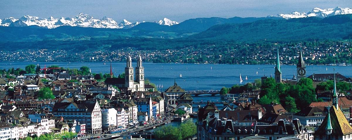 A view of Zurich, showing the city, the lake behind it, and behind that the snow-capped mountains of the Alps.