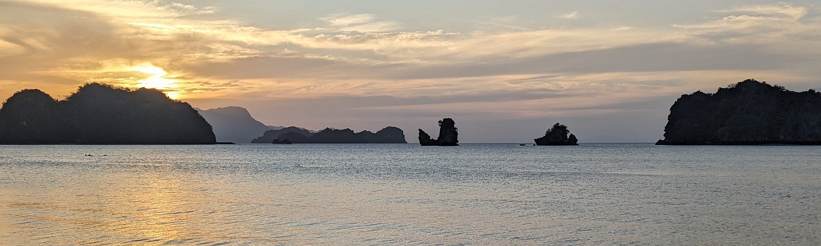 A beach with many rocky mountains sticking out from the the water, with the sun setting in the distance.