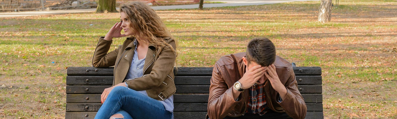 A man and woman sit on a park bench; she’s turned away from him looking into the distance and he is looking down, elbows resting on his knees and his hands are on is head.