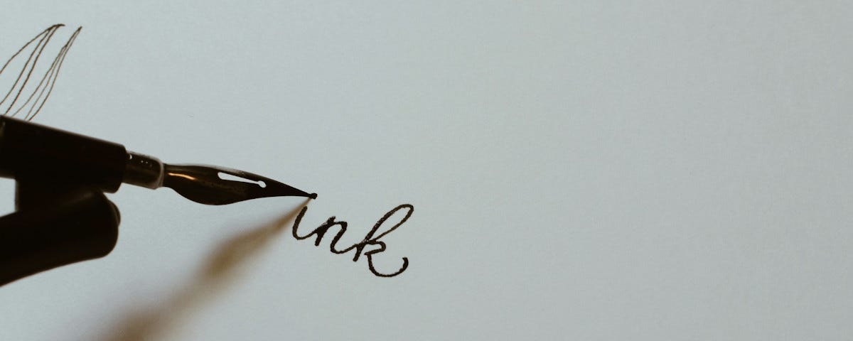 A fountain pen dotting the i of the word ‘ink’, which is written in neat copperplate in the centre of a pristine sheet of white paper.