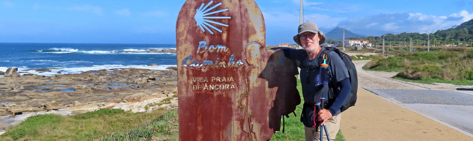 Man wearing a cap, backpack and carrying hiking poles leans against a rusted metal sign with a cutout of the Camino scallop shell and the words Bom Cominho and Vila Praia de Ancora. (Good Journey and Village Beach of Ancora)