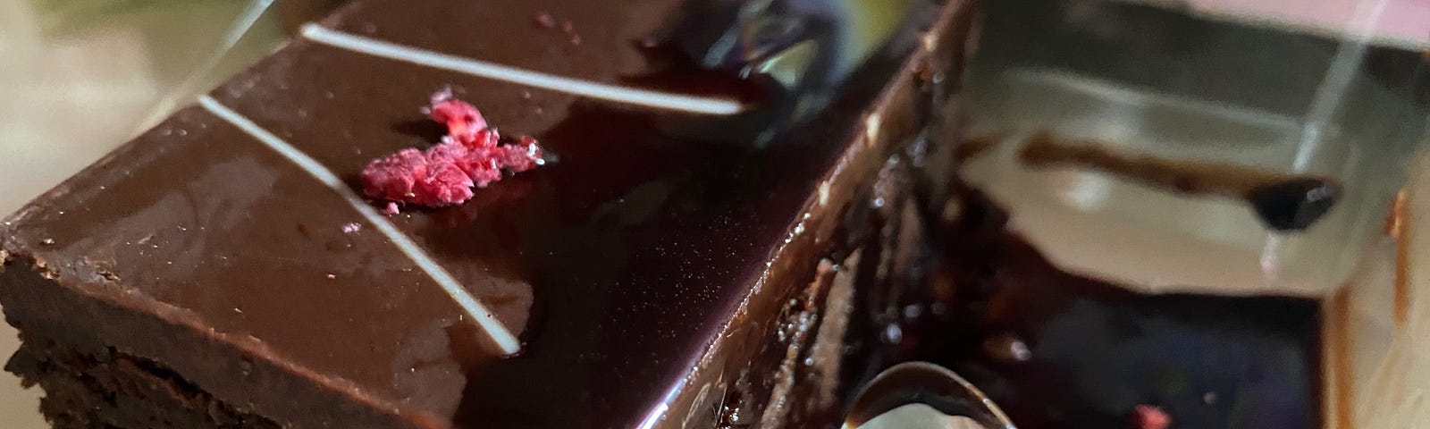 A rectangular portion of chocolate cake with a thick layer of chocolate on top and chocolate sauce. Part of a spoon is on the right of the cake.